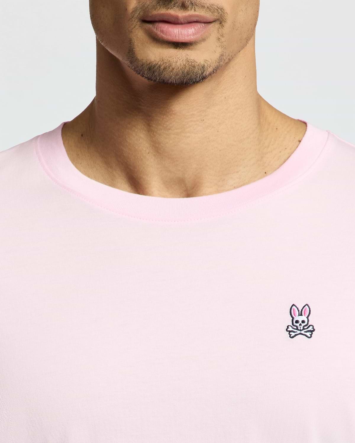 A close-up captures a person wearing the Psycho Bunny MENS CLASSIC CREW NECK TEE - B6U014CRPC in light pink. Made from soft pima cotton, it sports a small embroidered bunny with crossbones logo on the left. The regular fit tee shows their face partially from the nose down against a plain background.