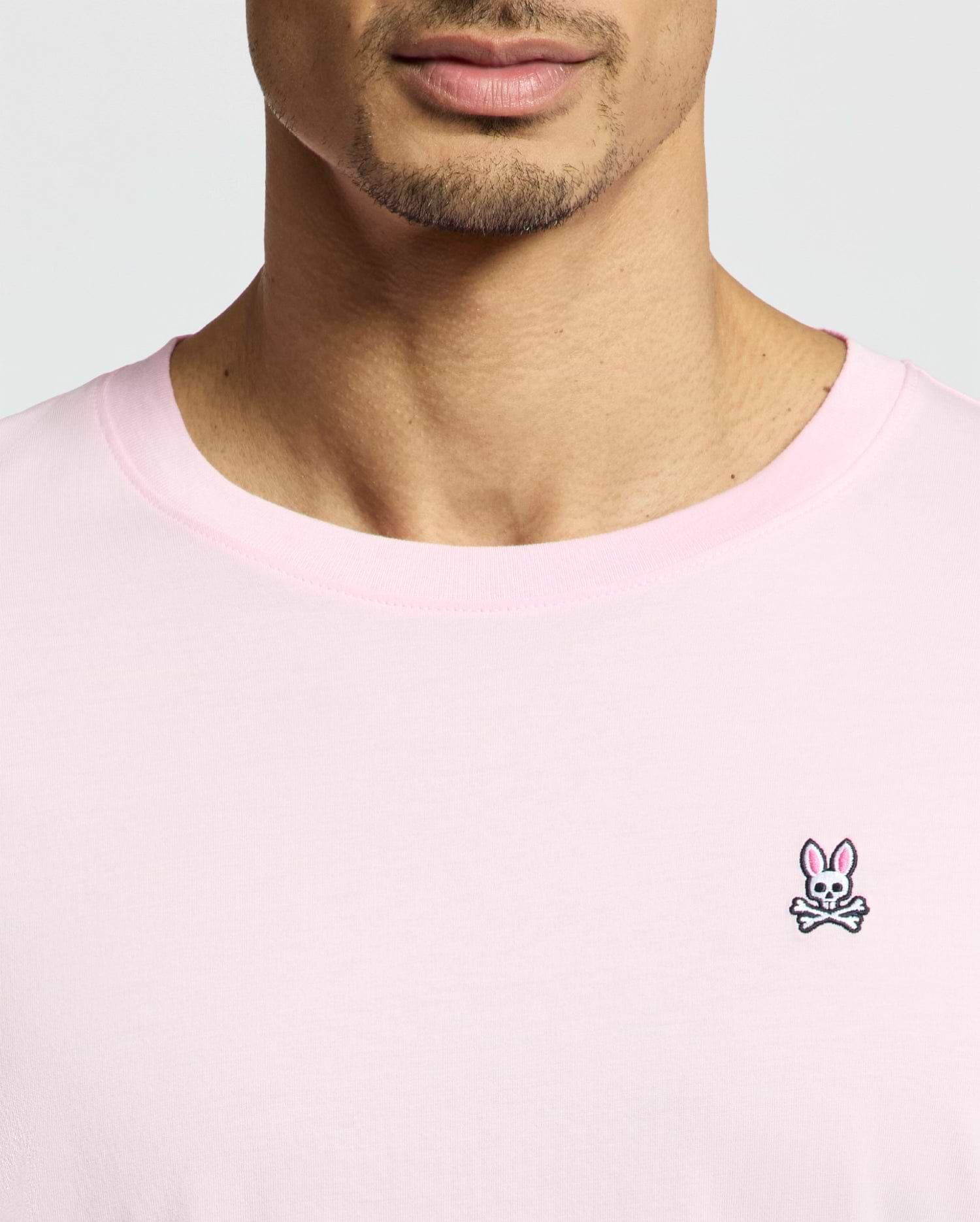 A close-up captures a person wearing the Psycho Bunny MENS CLASSIC CREW NECK TEE - B6U014CRPC in light pink. Made from soft pima cotton, it sports a small embroidered bunny with crossbones logo on the left. The regular fit tee shows their face partially from the nose down against a plain background.