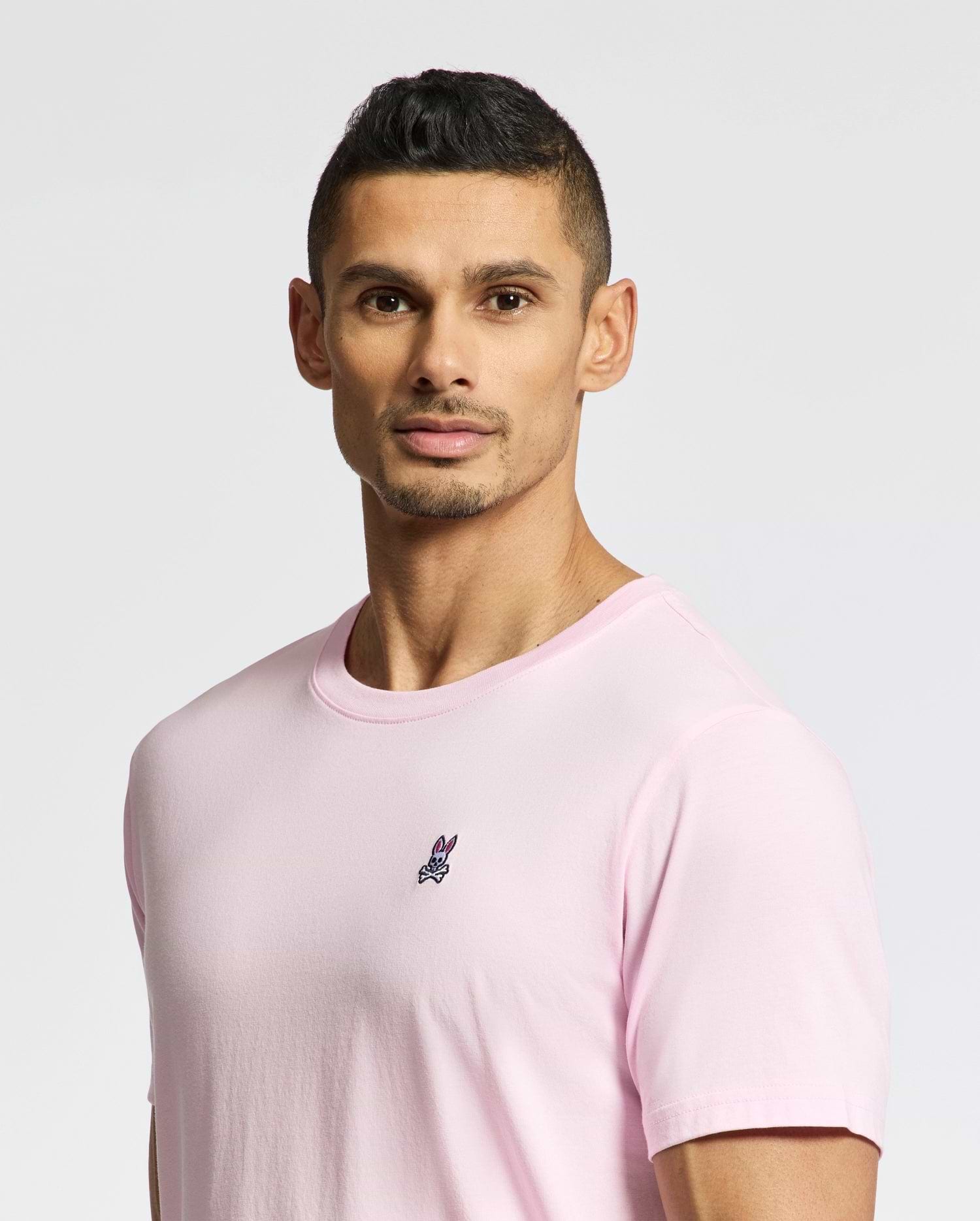 A man with short black hair and a trimmed beard wears the Psycho Bunny MENS CLASSIC CREW NECK TEE - B6U014CRPC in light pink, made from soft pima cotton with a small embroidered logo on the chest. He stands against a plain, light-toned background.
