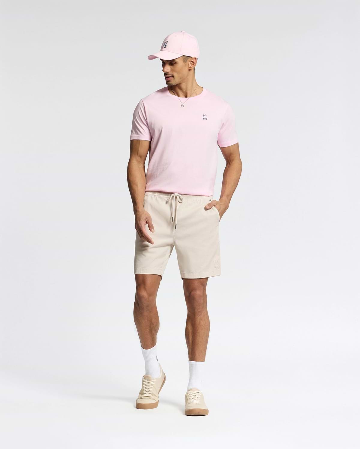 A man wears a light pink Psycho Bunny pima cotton cap and matching MENS CLASSIC CREW NECK TEE - B6U014CRPC, paired with beige drawstring shorts, white socks, and beige sneakers. Against a plain background, he looks down with his hands at his sides.