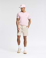 A man wears a light pink Psycho Bunny pima cotton cap and matching MENS CLASSIC CREW NECK TEE - B6U014CRPC, paired with beige drawstring shorts, white socks, and beige sneakers. Against a plain background, he looks down with his hands at his sides.