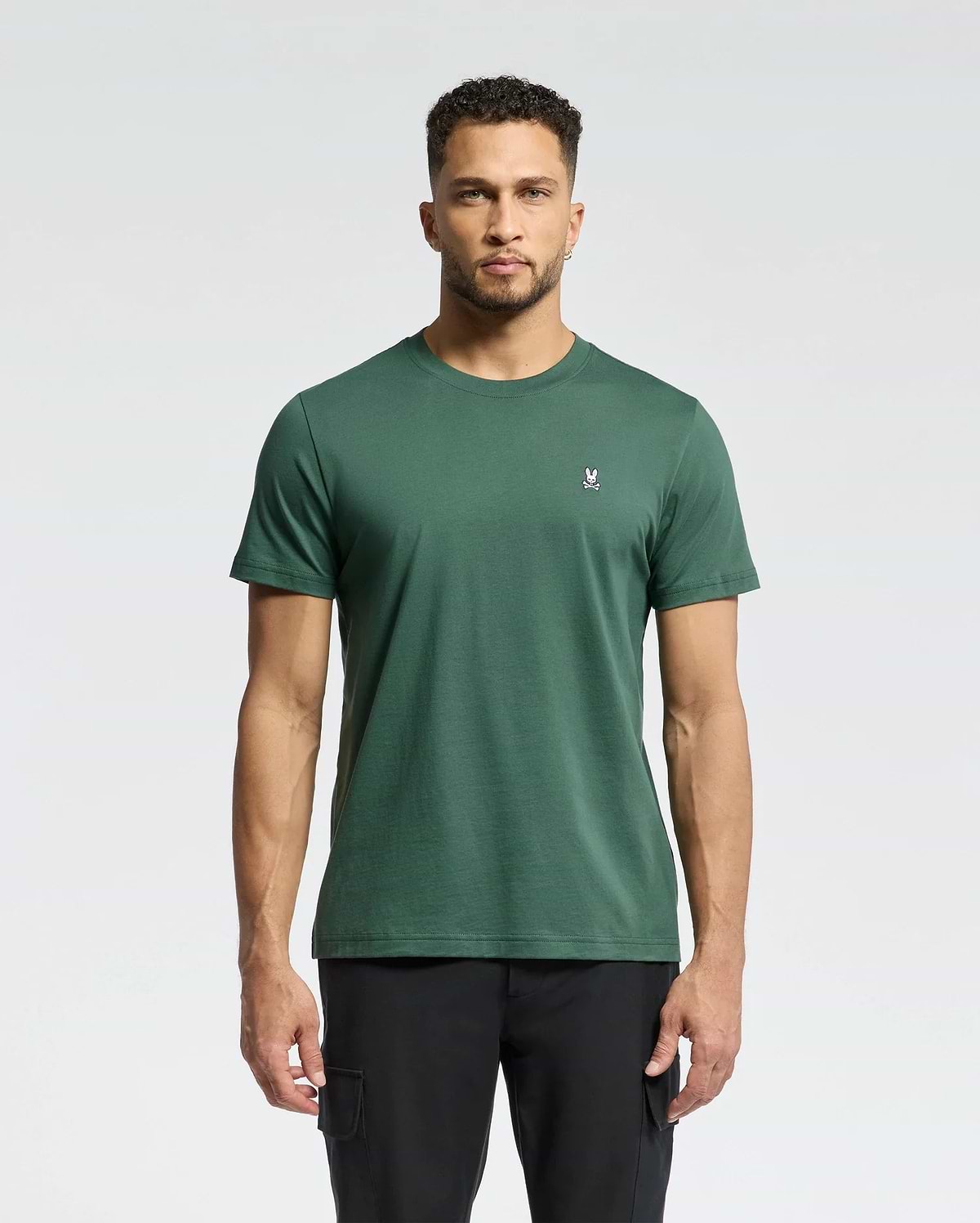 A man faces forward against a light gray background, wearing Psycho Bunny's MENS CLASSIC CREW NECK TEE - B6U014F200 in dark green with a small white rabbit logo on the left chest, paired with black pants.