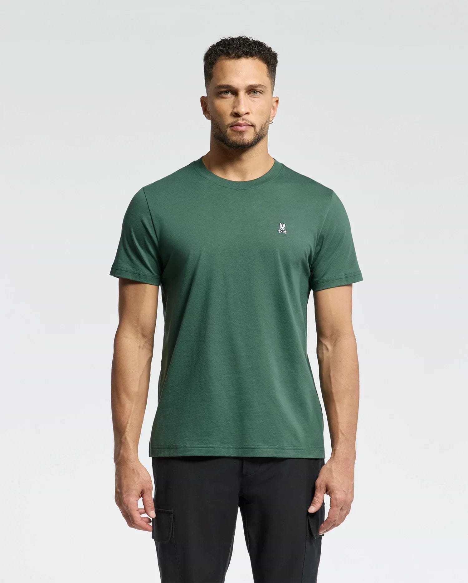 A man faces forward against a light gray background, wearing Psycho Bunny's MENS CLASSIC CREW NECK TEE - B6U014F200 in dark green with a small white rabbit logo on the left chest, paired with black pants.