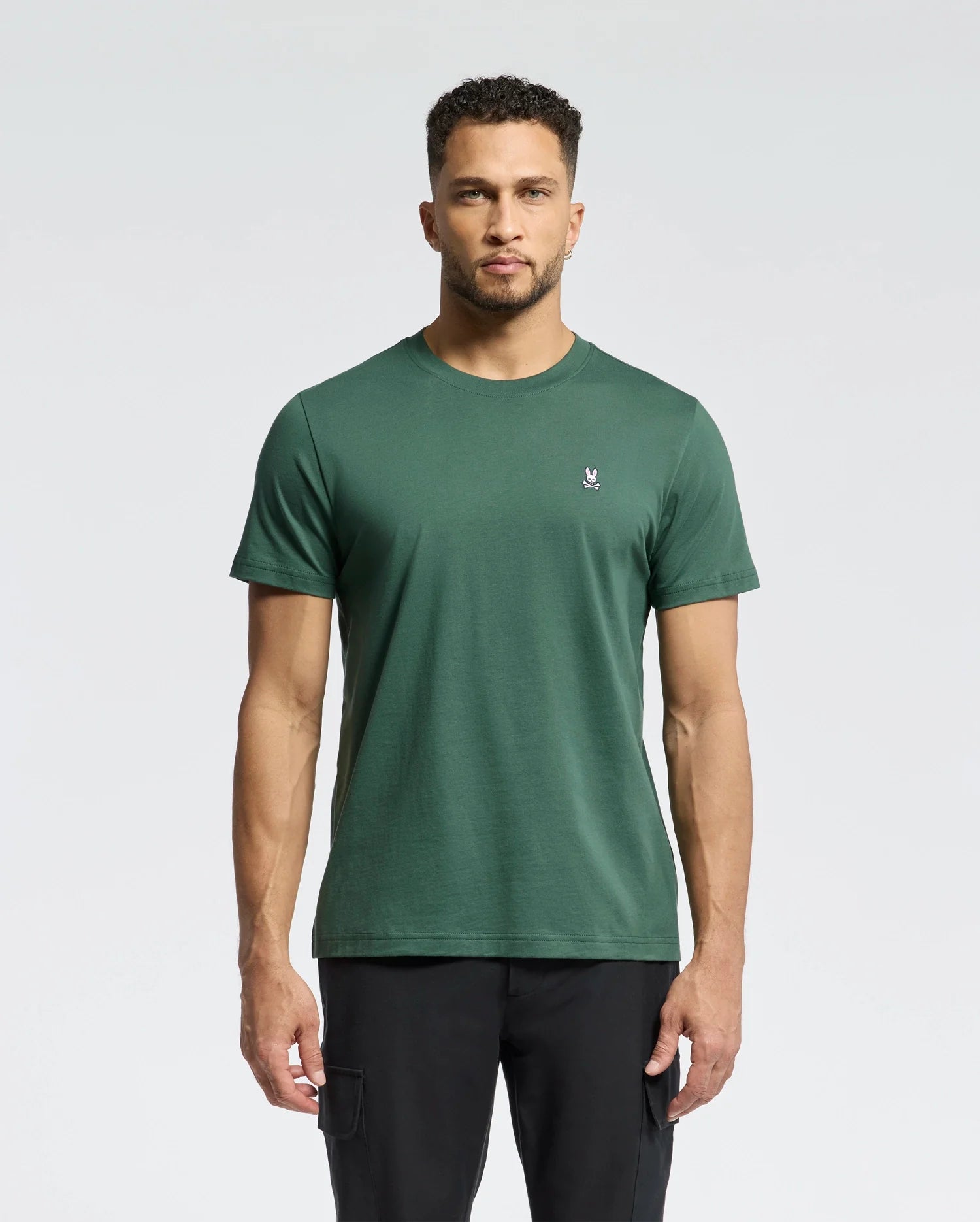 A man faces forward against a light gray background, wearing Psycho Bunny's MENS CLASSIC CREW NECK TEE - B6U014F200 in dark green with a small white rabbit logo on the left chest, paired with black pants.