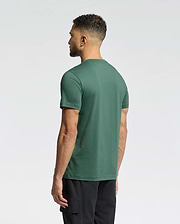 A man with short curly hair, wearing a green Psycho Bunny MENS CLASSIC CREW NECK TEE (B6U014F200) and black pants, stands with his back to the camera against a plain light background.