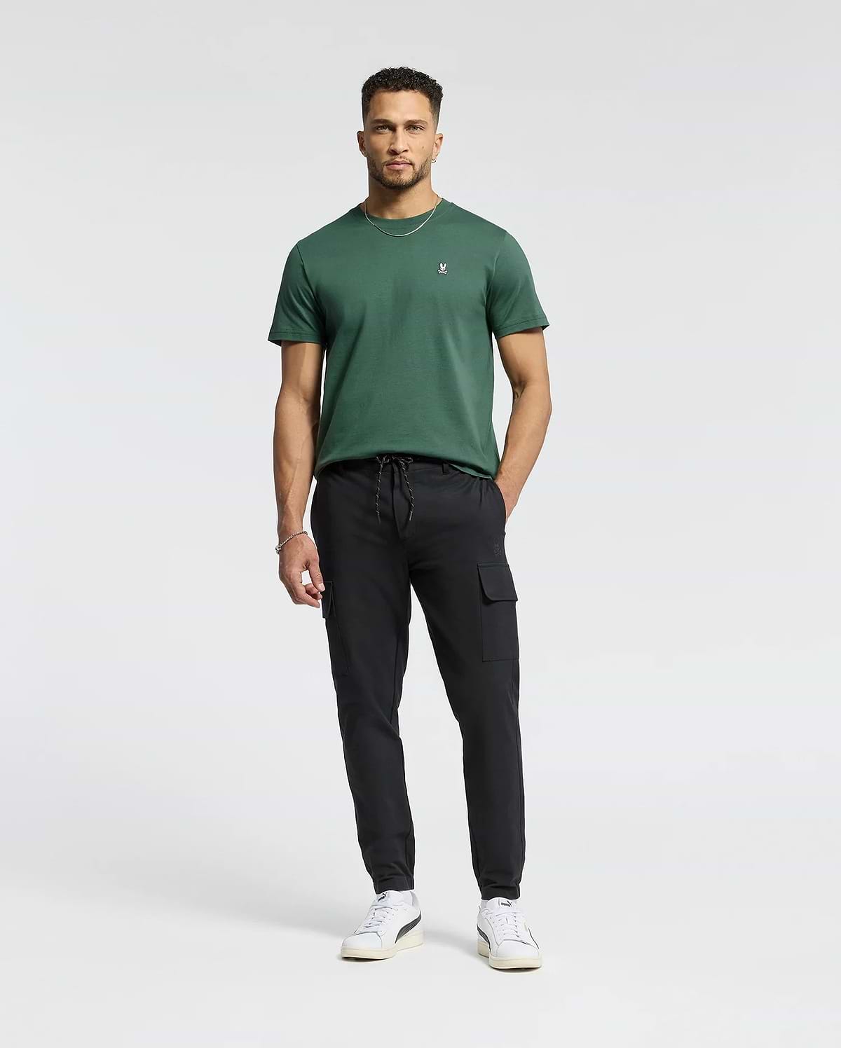 A man with short hair and a trimmed beard stands against a white background, wearing Psycho Bunny's MENS CLASSIC CREW NECK TEE - B6U014F200 in dark green, paired with black cargo pants and white sneakers, hands relaxed by his sides.