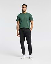 A man with short hair and a trimmed beard stands against a white background, wearing Psycho Bunny's MENS CLASSIC CREW NECK TEE - B6U014F200 in dark green, paired with black cargo pants and white sneakers, hands relaxed by his sides.