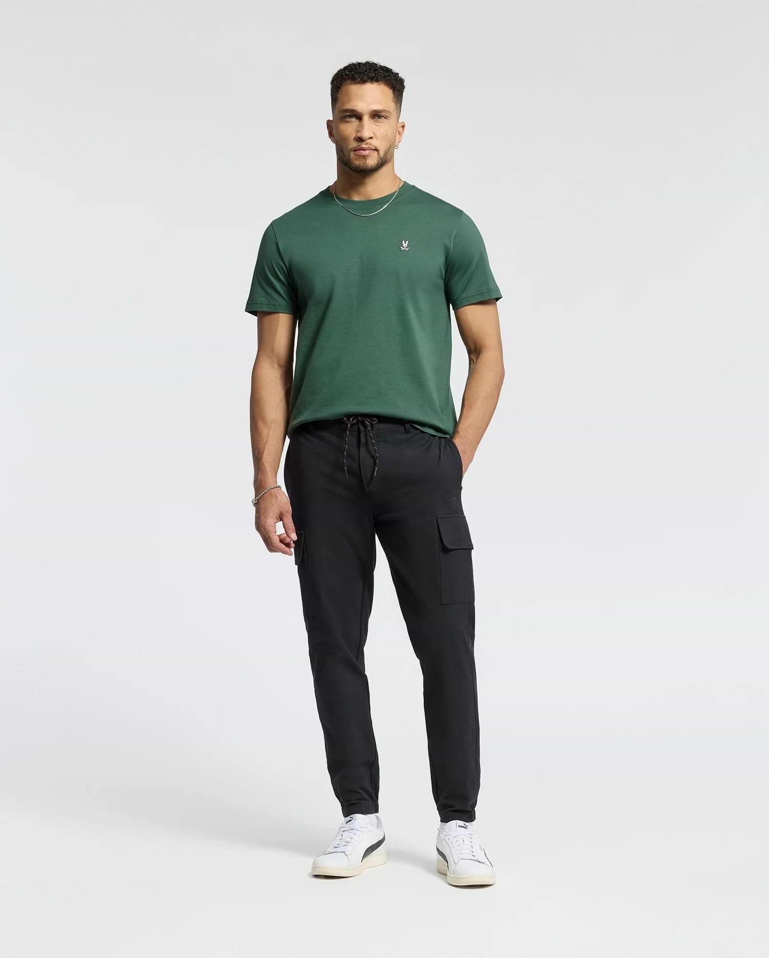 A man with short hair and a trimmed beard stands against a white background, wearing Psycho Bunny's MENS CLASSIC CREW NECK TEE - B6U014F200 in dark green, paired with black cargo pants and white sneakers, hands relaxed by his sides.