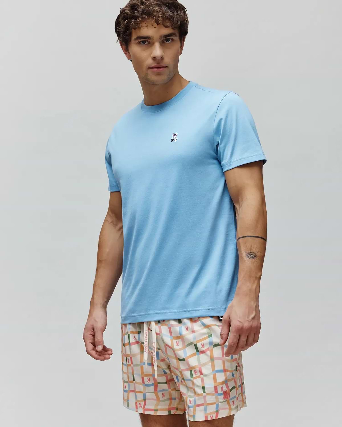Mens Classic Crew Neck Tee in light blue premium jersey, featuring a classic fit and a small, multi-color embroidered Psycho Bunny skull logo on the left chest, paired with cream shorts featuring a colorful grid pattern and small bunny logos. (2)