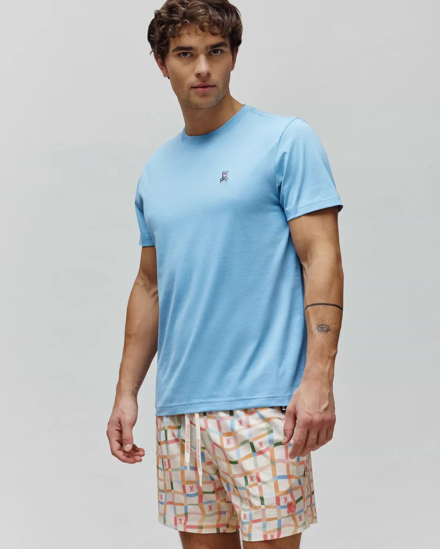 Mens Classic Crew Neck Tee in light blue premium jersey, featuring a classic fit and a small, multi-color embroidered Psycho Bunny skull logo on the left chest, paired with cream shorts featuring a colorful grid pattern and small bunny logos. (2)