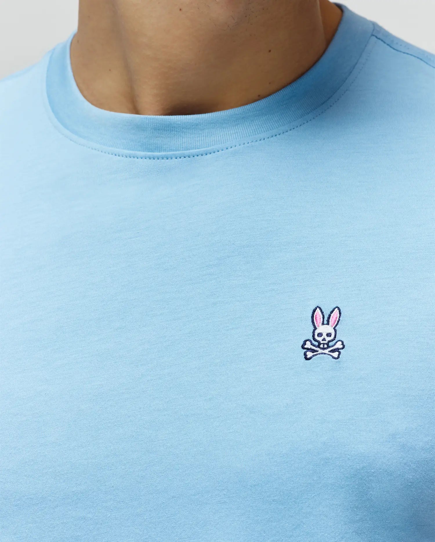 Light blue Mens Classic Crew Neck Tee, crafted from soft jersey fabric, featuring a small, intricately embroidered Psycho Bunny skull and crossbones logo with pink and white accents on the left chest. (3)