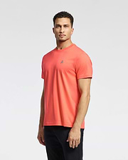 A man in the Psycho Bunny MENS CLASSIC CREW NECK TEE - B6U014F200, featuring a coral color and small embroidered Bunny logo, stands against a white background. He has short curly hair, a serious expression, and pairs the tee with black pants.