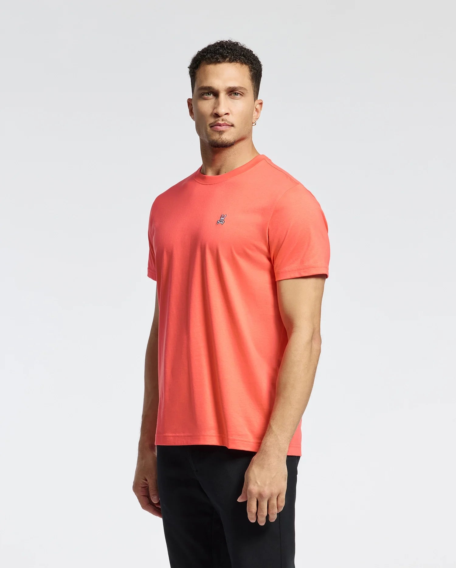 A man in the Psycho Bunny MENS CLASSIC CREW NECK TEE - B6U014F200, featuring a coral color and small embroidered Bunny logo, stands against a white background. He has short curly hair, a serious expression, and pairs the tee with black pants.