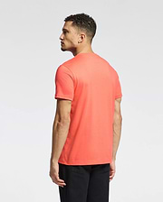 A person with short curly hair wears a Psycho Bunny MENS CLASSIC CREW NECK TEE in bright coral, featuring an embroidered Bunny logo, and black pants. They face away from the camera against a plain white background.