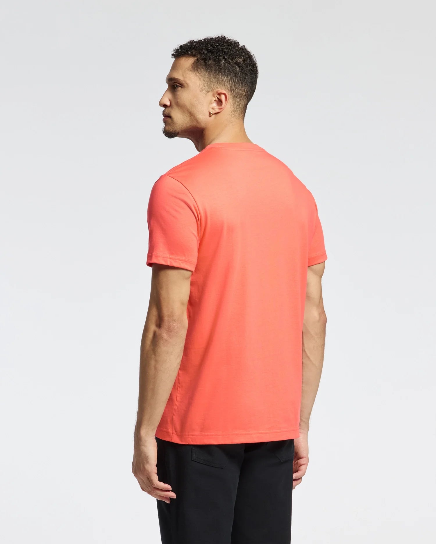A person with short curly hair wears a Psycho Bunny MENS CLASSIC CREW NECK TEE in bright coral, featuring an embroidered Bunny logo, and black pants. They face away from the camera against a plain white background.