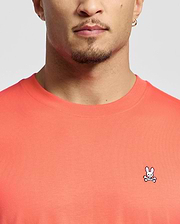 A person is wearing the Psycho Bunny MENS CLASSIC CREW NECK TEE in coral, made of soft Pima cotton. It features an embroidered Bunny logo above crossbones on the chest, with only a glimpse of their face visible.