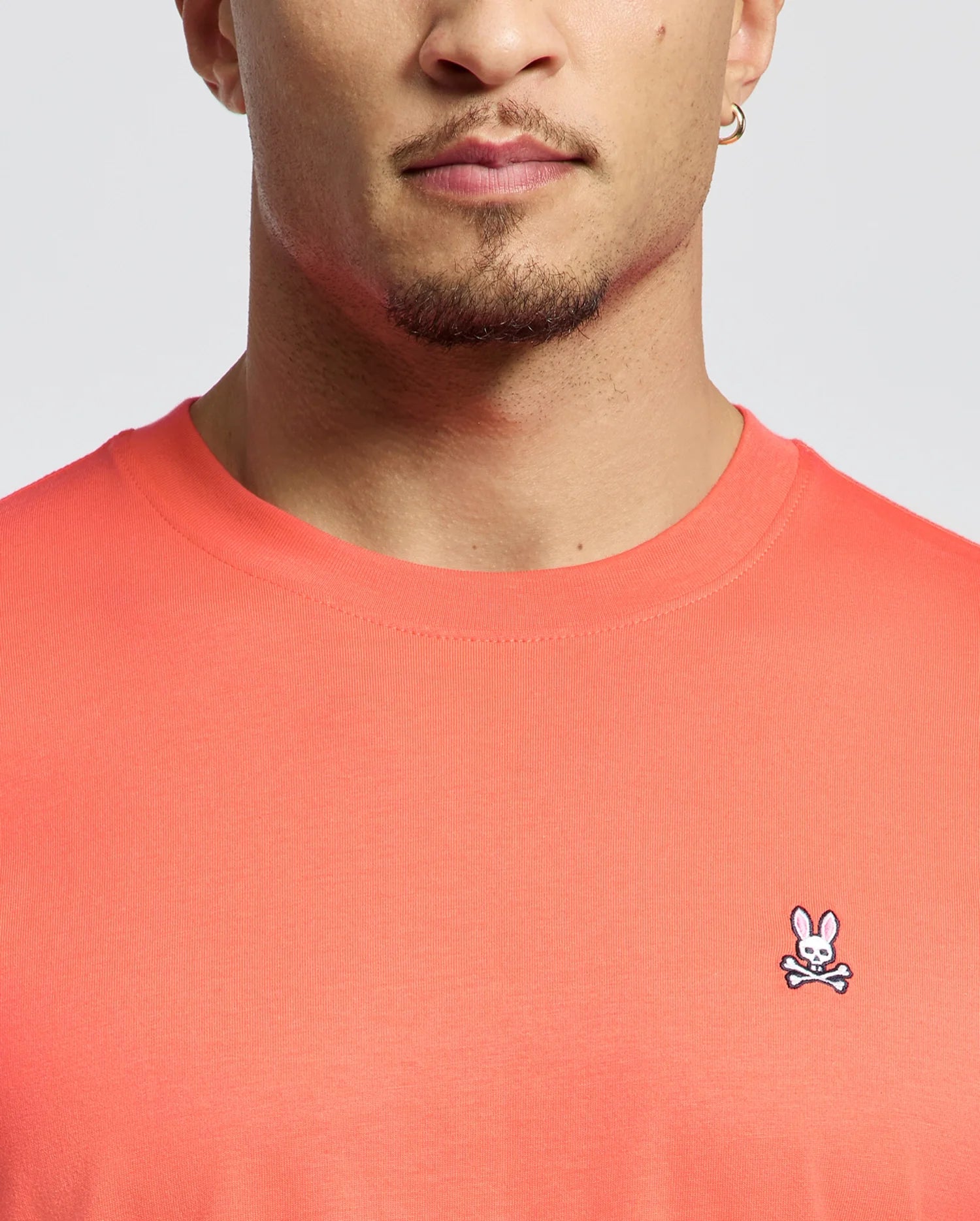 A person is wearing the Psycho Bunny MENS CLASSIC CREW NECK TEE in coral, made of soft Pima cotton. It features an embroidered Bunny logo above crossbones on the chest, with only a glimpse of their face visible.