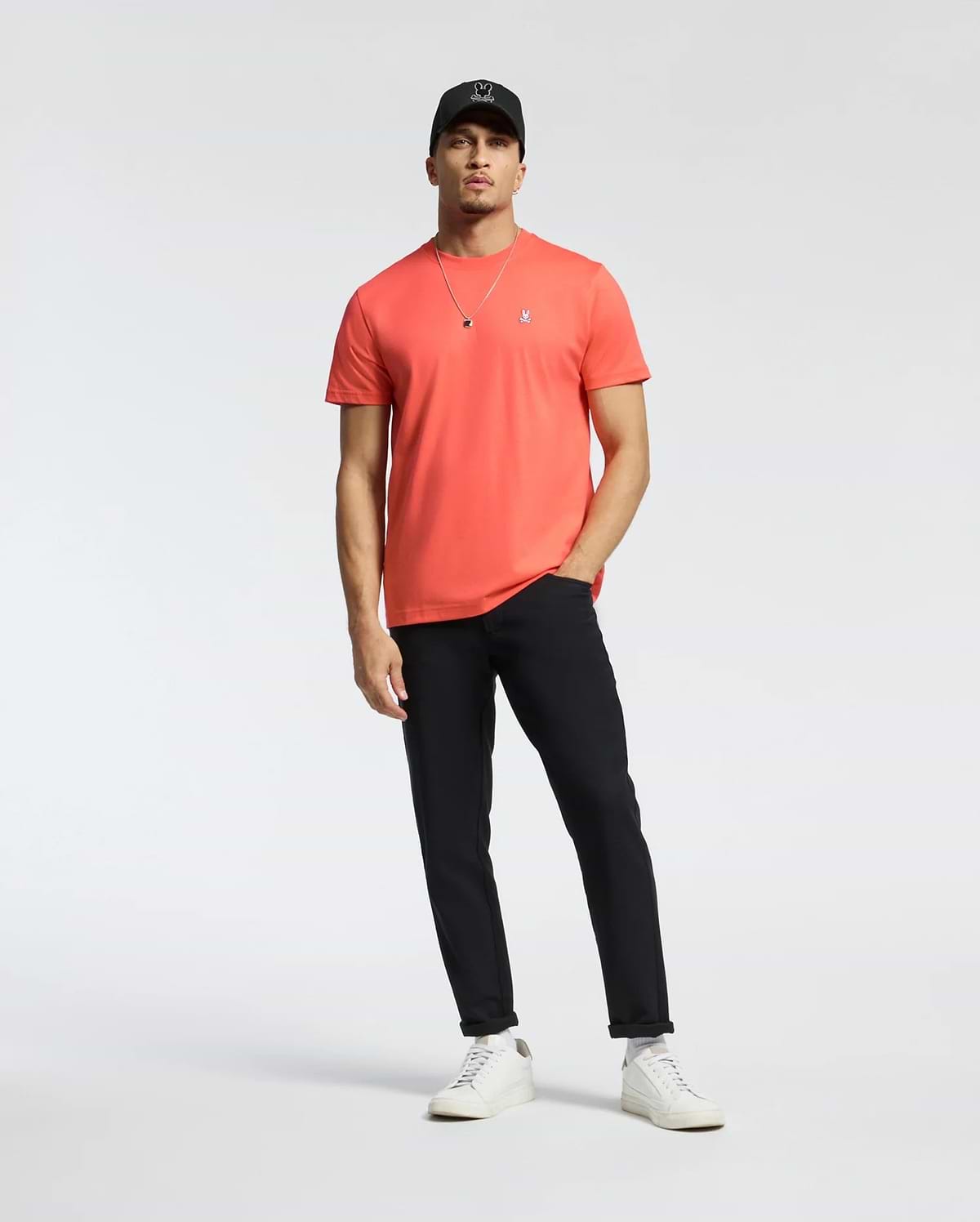 A man in a coral Psycho Bunny MENS CLASSIC CREW NECK TEE (B6U014F200) with an embroidered Bunny logo, black pants, white sneakers, and a black cap stands relaxed against a plain white background. One hand rests in his pocket as he wears a necklace.