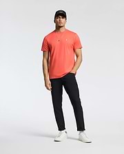 A man in a coral Psycho Bunny MENS CLASSIC CREW NECK TEE (B6U014F200) with an embroidered Bunny logo, black pants, white sneakers, and a black cap stands relaxed against a plain white background. One hand rests in his pocket as he wears a necklace.
