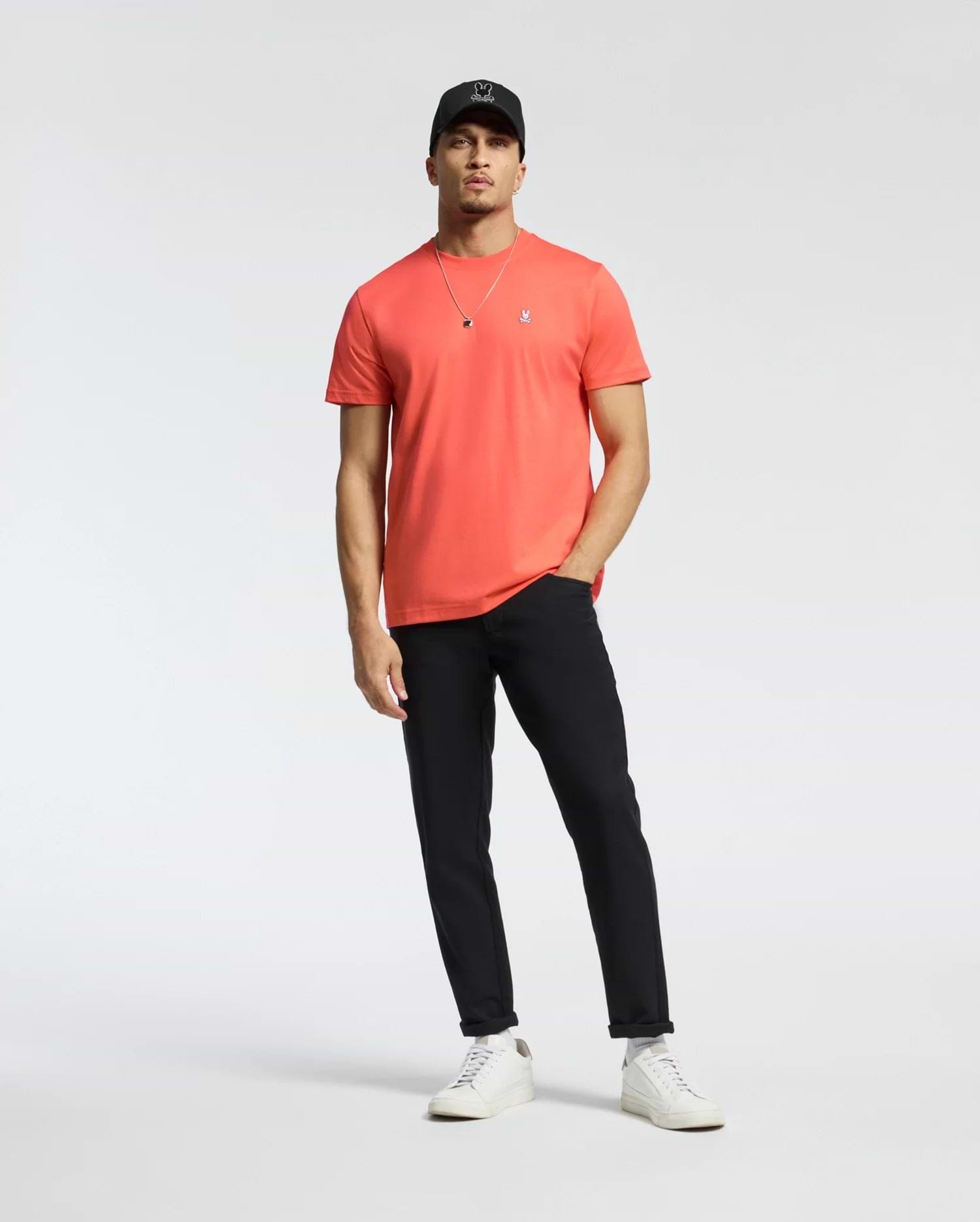 A man in a coral Psycho Bunny MENS CLASSIC CREW NECK TEE (B6U014F200) with an embroidered Bunny logo, black pants, white sneakers, and a black cap stands relaxed against a plain white background. One hand rests in his pocket as he wears a necklace.