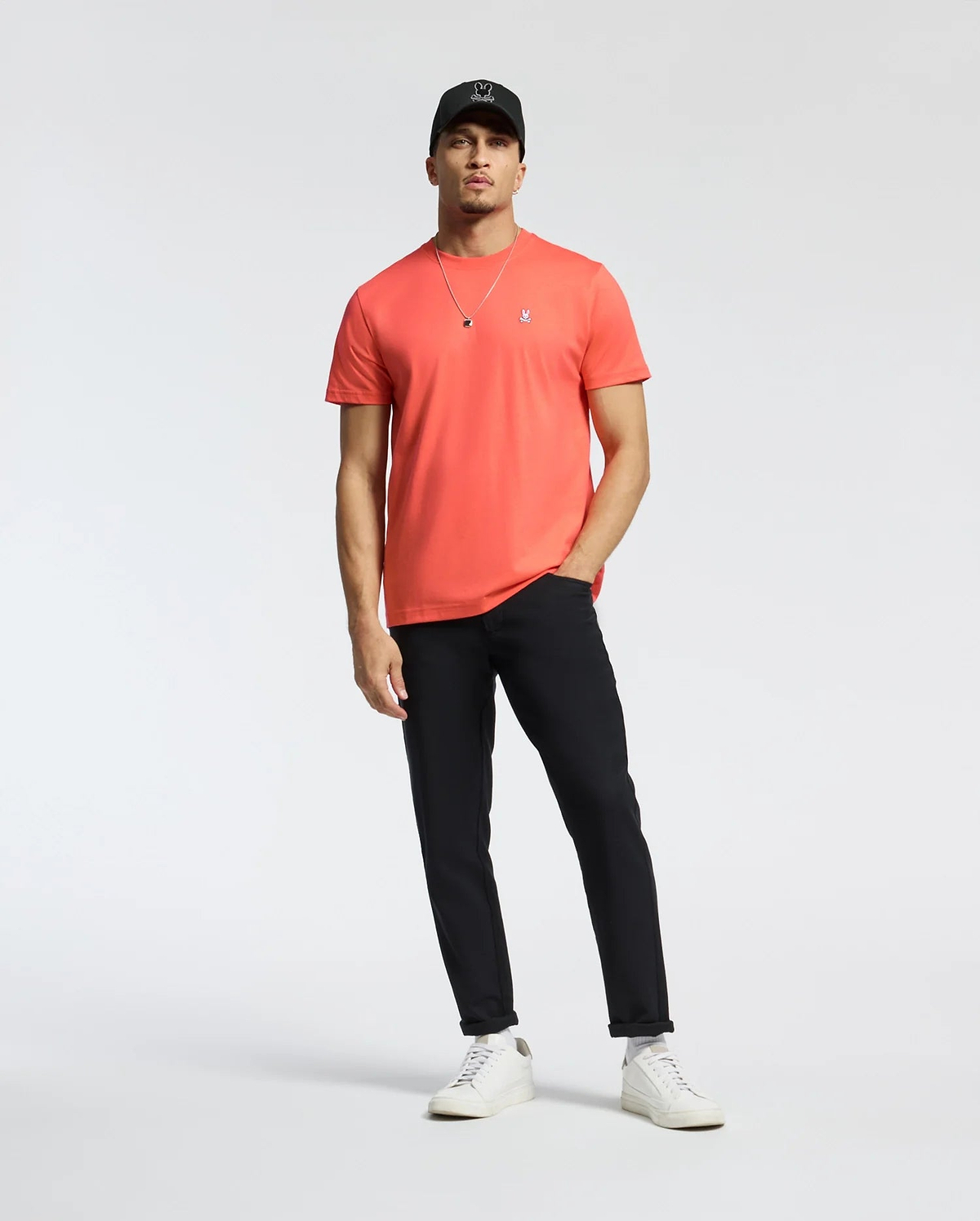 A man in a coral Psycho Bunny MENS CLASSIC CREW NECK TEE (B6U014F200) with an embroidered Bunny logo, black pants, white sneakers, and a black cap stands relaxed against a plain white background. One hand rests in his pocket as he wears a necklace.