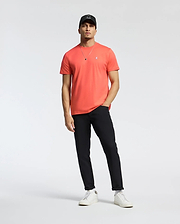 A man in a coral Psycho Bunny MENS CLASSIC CREW NECK TEE (B6U014F200) with an embroidered Bunny logo, black pants, white sneakers, and a black cap stands relaxed against a plain white background. One hand rests in his pocket as he wears a necklace.