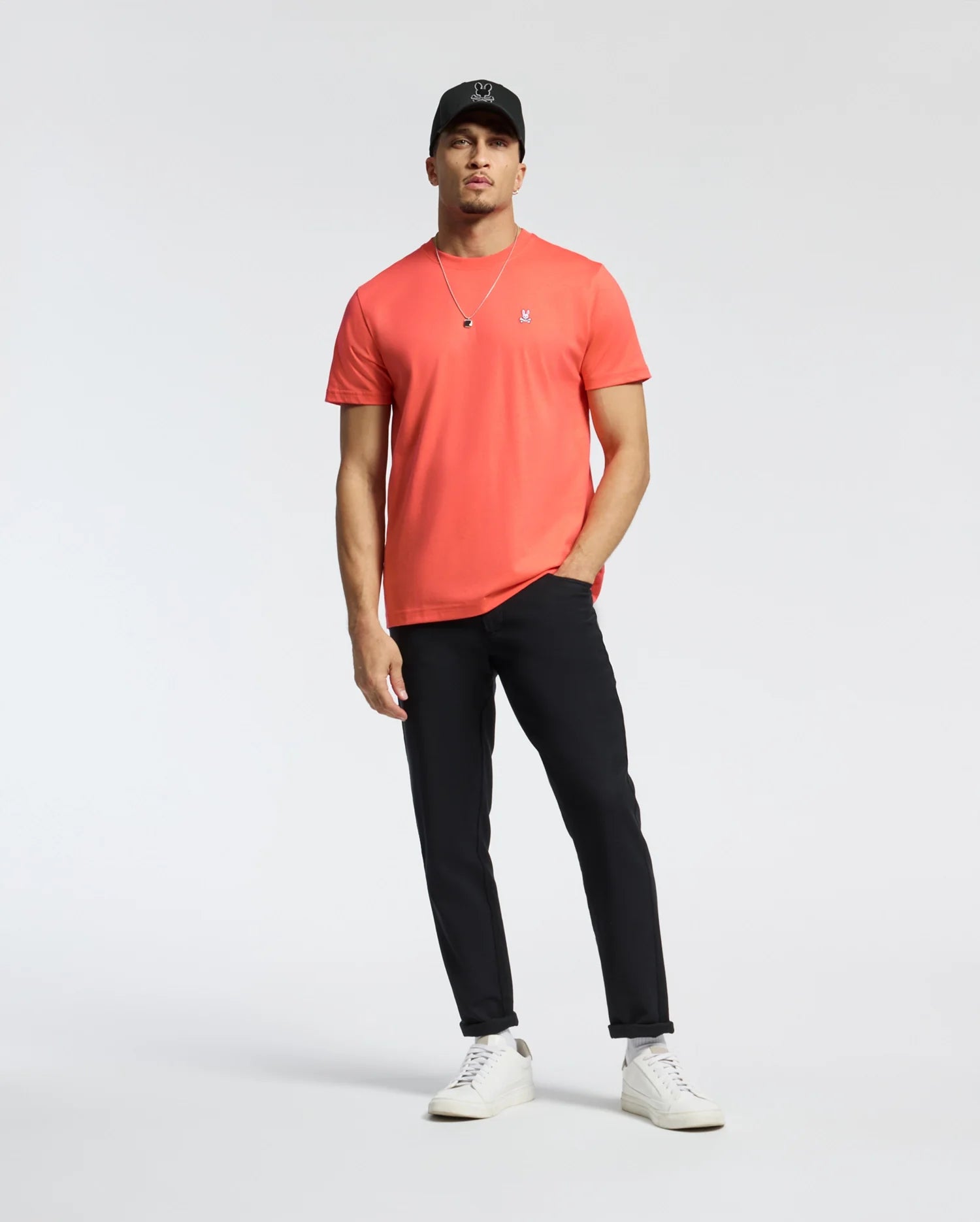 A man in a coral Psycho Bunny MENS CLASSIC CREW NECK TEE (B6U014F200) with an embroidered Bunny logo, black pants, white sneakers, and a black cap stands relaxed against a plain white background. One hand rests in his pocket as he wears a necklace.