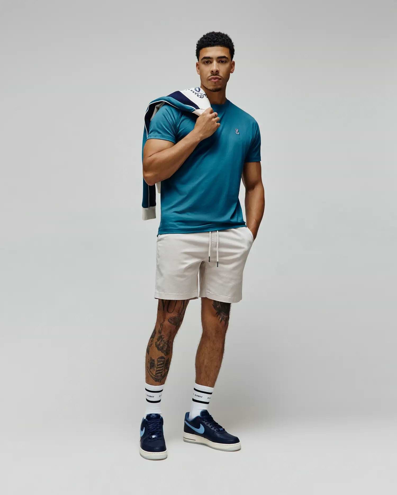 Mens Classic Crew Neck Tee in teal jersey with a small embroidered Psycho Bunny skull logo on the left chest, paired with light beige drawstring shorts and a navy blue and white patterned sweatshirt draped over the shoulder. (4)