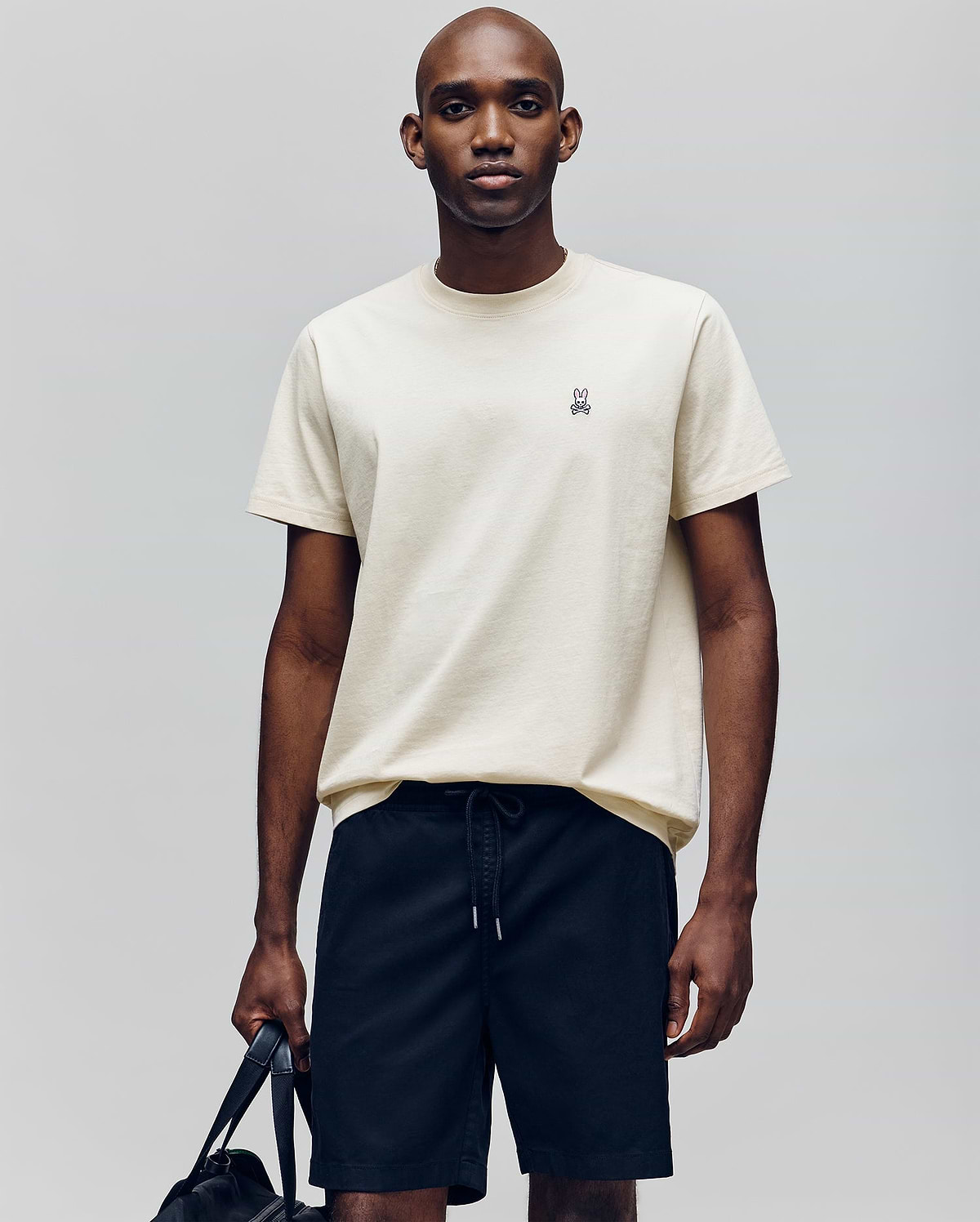 A man stands against a plain background wearing the Psycho Bunny MENS CLASSIC CREW NECK TEE - B6U014F200, a light beige, regular-fit pima cotton tee with an embroidered bunny logo on the chest, paired with black drawstring shorts and holding a black bag.