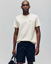 A man stands against a plain background wearing the Psycho Bunny MENS CLASSIC CREW NECK TEE - B6U014F200, a light beige, regular-fit pima cotton tee with an embroidered bunny logo on the chest, paired with black drawstring shorts and holding a black bag.