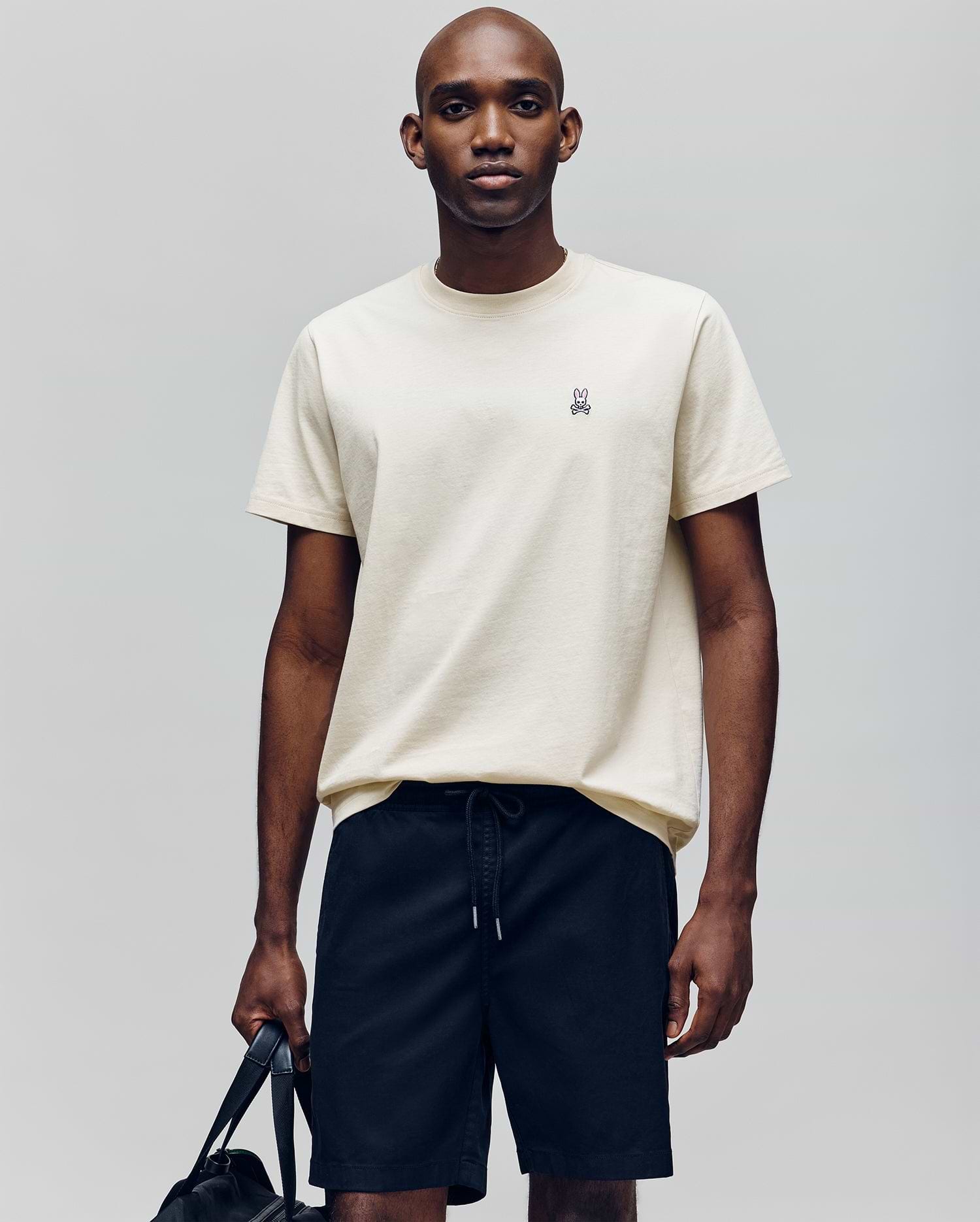 A man stands against a plain background wearing the Psycho Bunny MENS CLASSIC CREW NECK TEE - B6U014F200, a light beige, regular-fit pima cotton tee with an embroidered bunny logo on the chest, paired with black drawstring shorts and holding a black bag.
