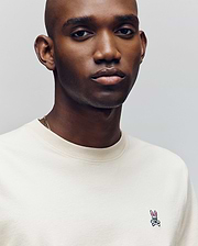 A man with a shaved head wears a cream Psycho Bunny MENS CLASSIC CREW NECK TEE (B6U014F200), featuring regular fit pima cotton and a small pink bunny and crossbones logo on the chest, looking at the camera with a neutral expression.