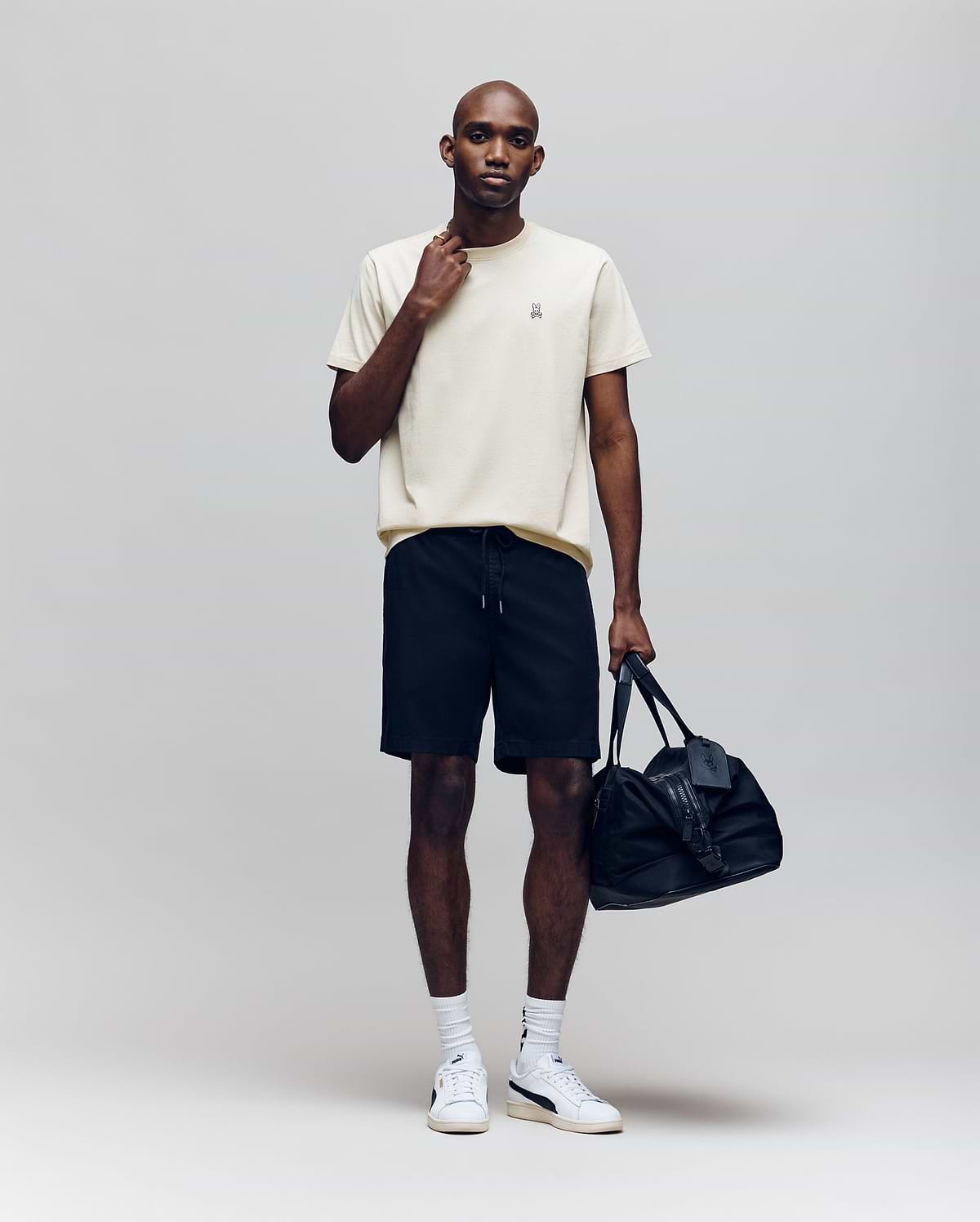 A man stands against a plain background wearing the Psycho Bunny MENS CLASSIC CREW NECK TEE - B6U014F200 in cream, paired with black shorts, white socks, white sneakers, holding a black duffel bag and touching his neck.
