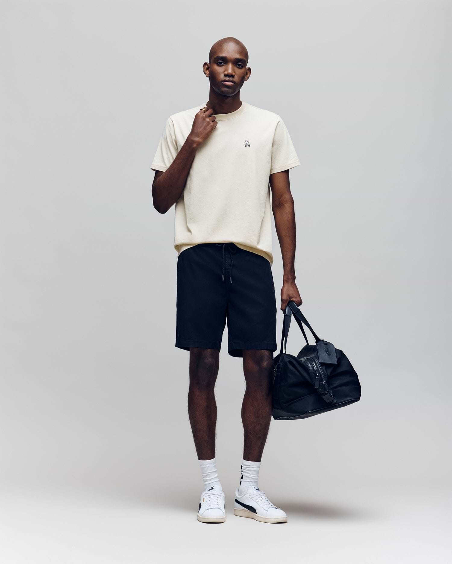 A man stands against a plain background wearing the Psycho Bunny MENS CLASSIC CREW NECK TEE - B6U014F200 in cream, paired with black shorts, white socks, white sneakers, holding a black duffel bag and touching his neck.