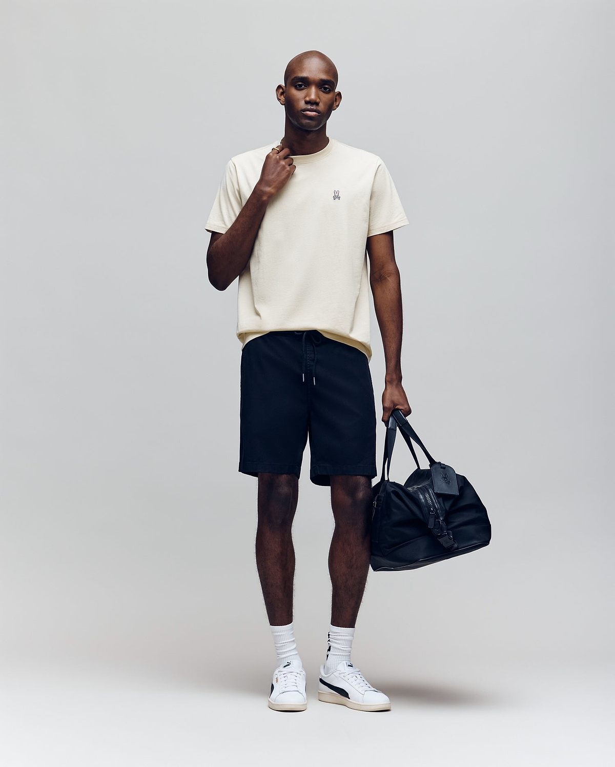 A man stands against a plain background wearing the Psycho Bunny MENS CLASSIC CREW NECK TEE - B6U014F200 in cream, paired with black shorts, white socks, white sneakers, holding a black duffel bag and touching his neck.