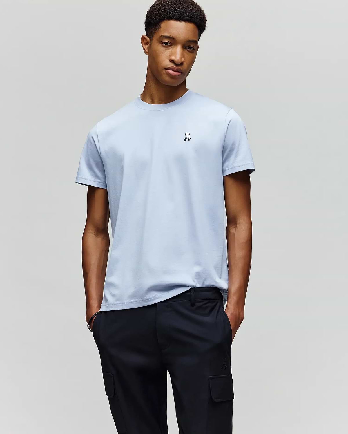 A man stands against a plain background, hands in pockets, wearing the Psycho Bunny MENS CLASSIC CREW NECK TEE - B6U014F200 in light blue Pima cotton with a small chest logo and dark cargo pants, looking relaxed.
