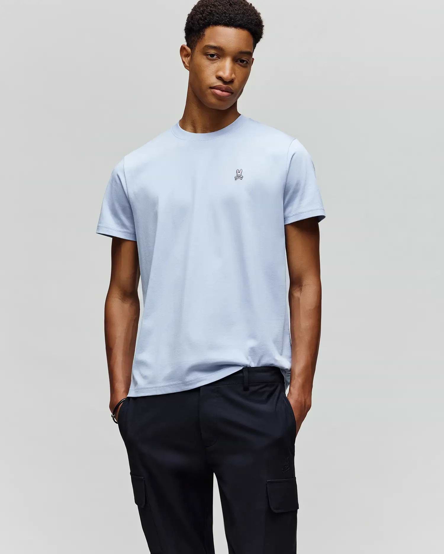 A man stands against a plain background, hands in pockets, wearing the Psycho Bunny MENS CLASSIC CREW NECK TEE - B6U014F200 in light blue Pima cotton with a small chest logo and dark cargo pants, looking relaxed.