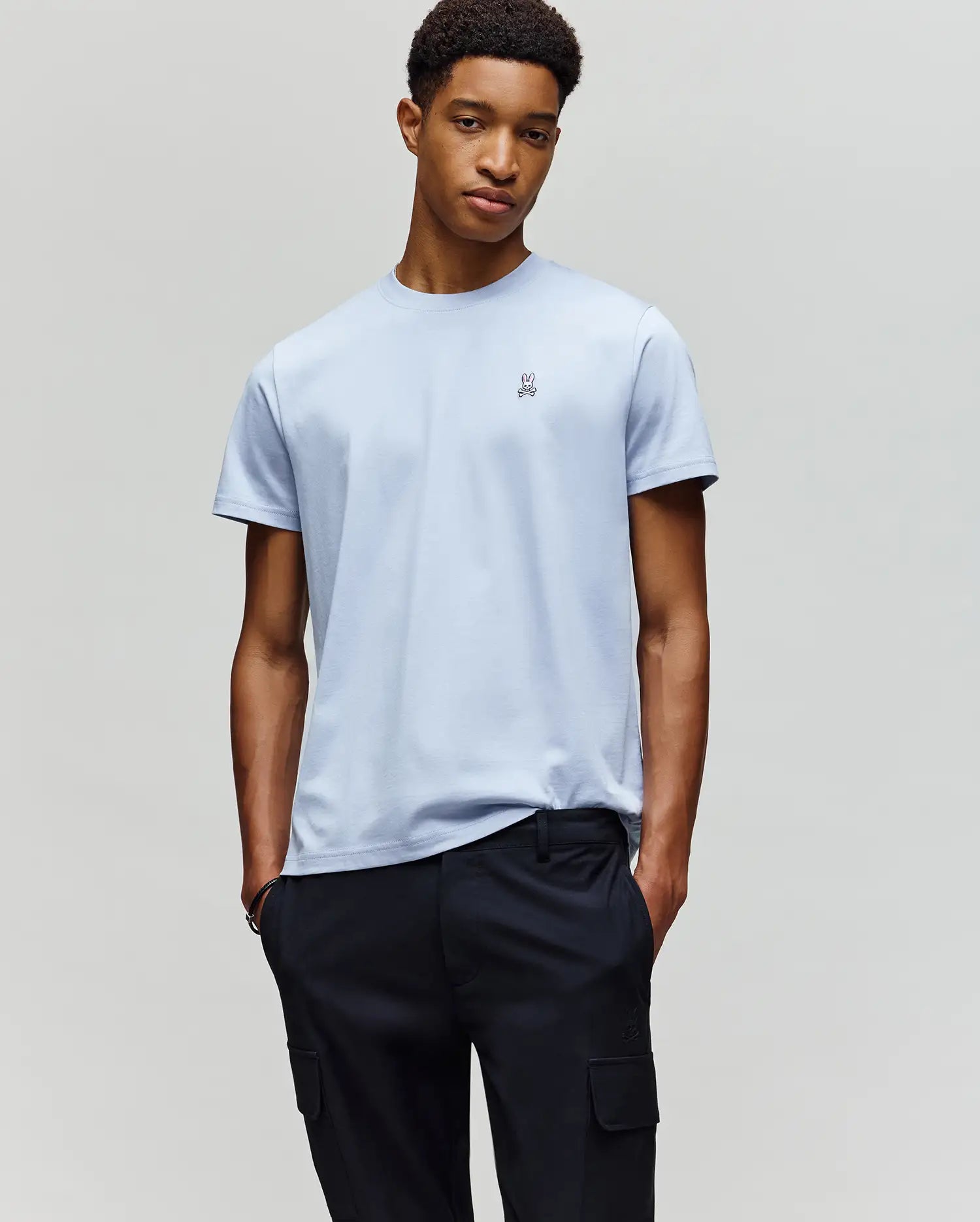 A man stands against a plain background, hands in pockets, wearing the Psycho Bunny MENS CLASSIC CREW NECK TEE - B6U014F200 in light blue Pima cotton with a small chest logo and dark cargo pants, looking relaxed.