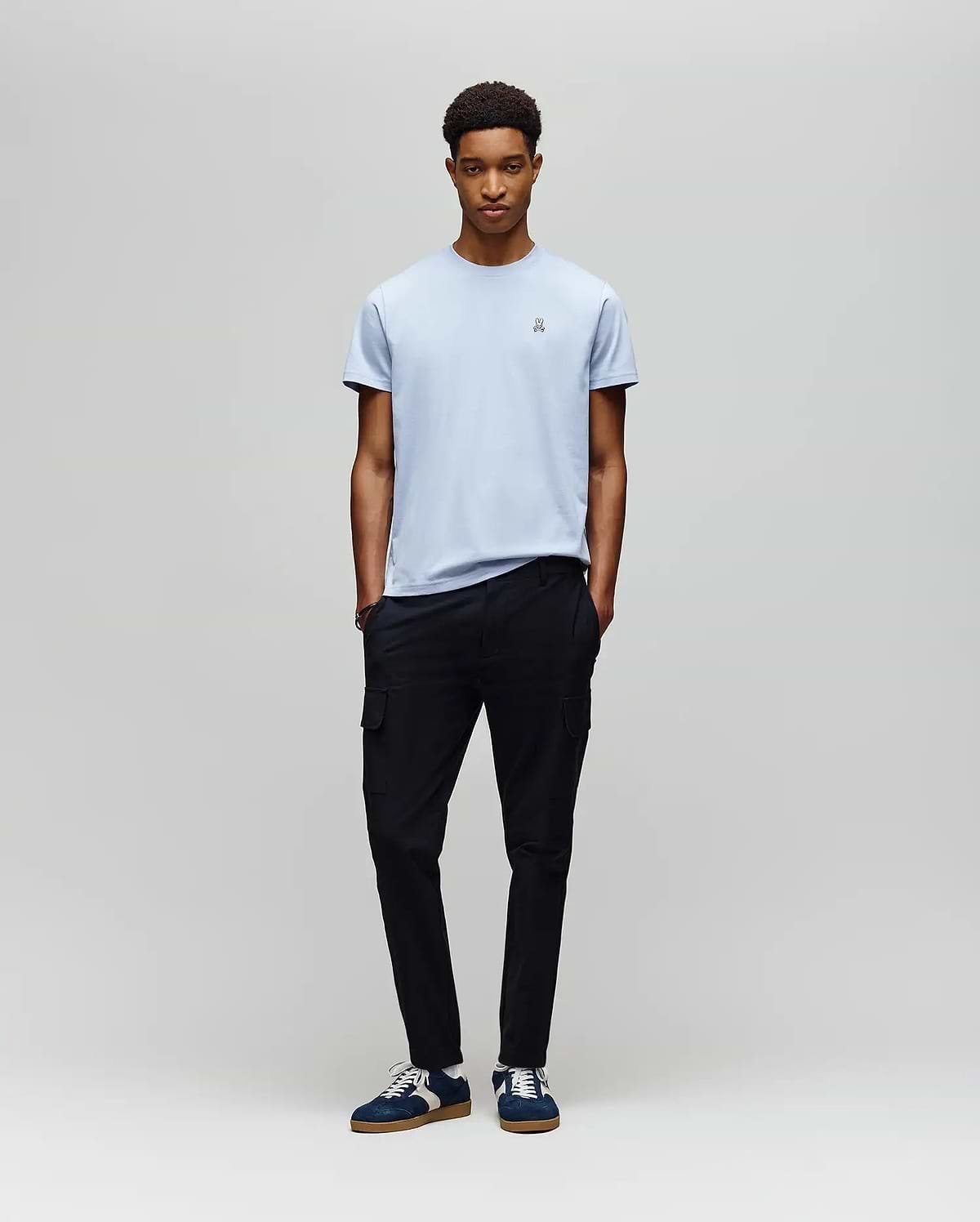 A young man with short curly hair stands against a light gray backdrop, hands in pockets, wearing a Psycho Bunny MENS CLASSIC CREW NECK TEE (B6U014F200) in light blue, black cargo pants, and navy sneakers with white laces.