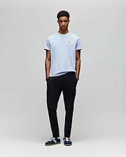 A young man with short curly hair stands against a light gray backdrop, hands in pockets, wearing a Psycho Bunny MENS CLASSIC CREW NECK TEE (B6U014F200) in light blue, black cargo pants, and navy sneakers with white laces.