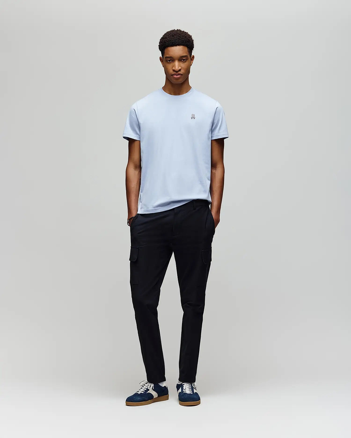 A young man with short curly hair stands against a light gray backdrop, hands in pockets, wearing a Psycho Bunny MENS CLASSIC CREW NECK TEE (B6U014F200) in light blue, black cargo pants, and navy sneakers with white laces.