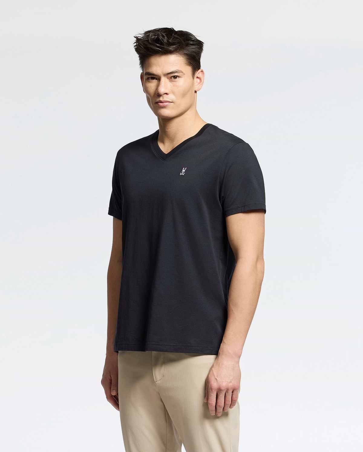 A person with short dark hair wearing a Psycho Bunny Men's Classic V Neck Tee (B6U100ARPC) in black and beige pants stands against a plain white background.