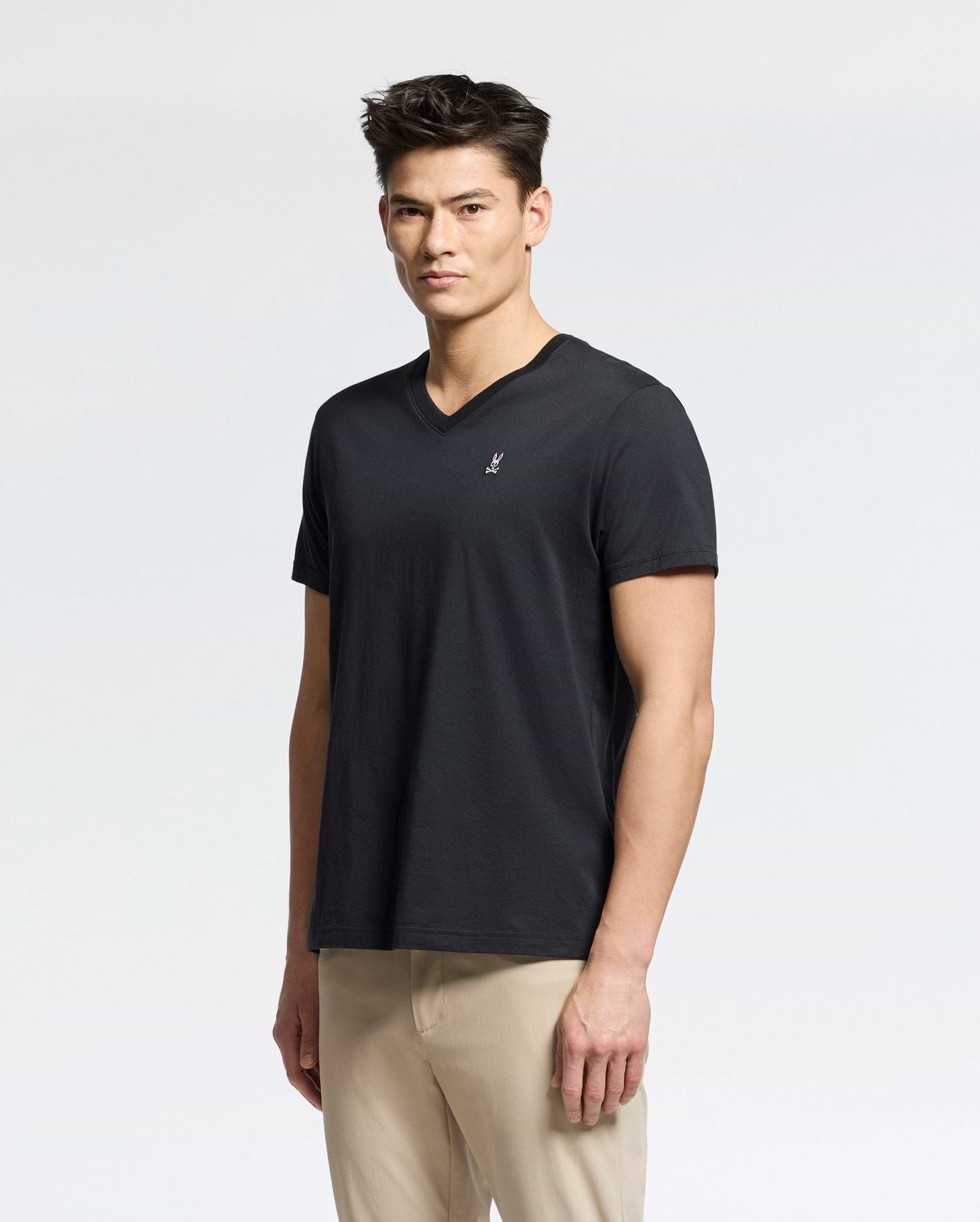 A person with short dark hair wearing a Psycho Bunny Men's Classic V Neck Tee (B6U100ARPC) in black and beige pants stands against a plain white background.