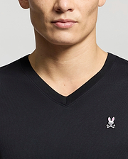 A person is wearing a Psycho Bunny Men's Classic V-Neck Tee (B6U100ARPC) in black, showcasing the pink bunny head and crossbones logo on the left side. The image is cropped to display only the upper torso and part of the face.