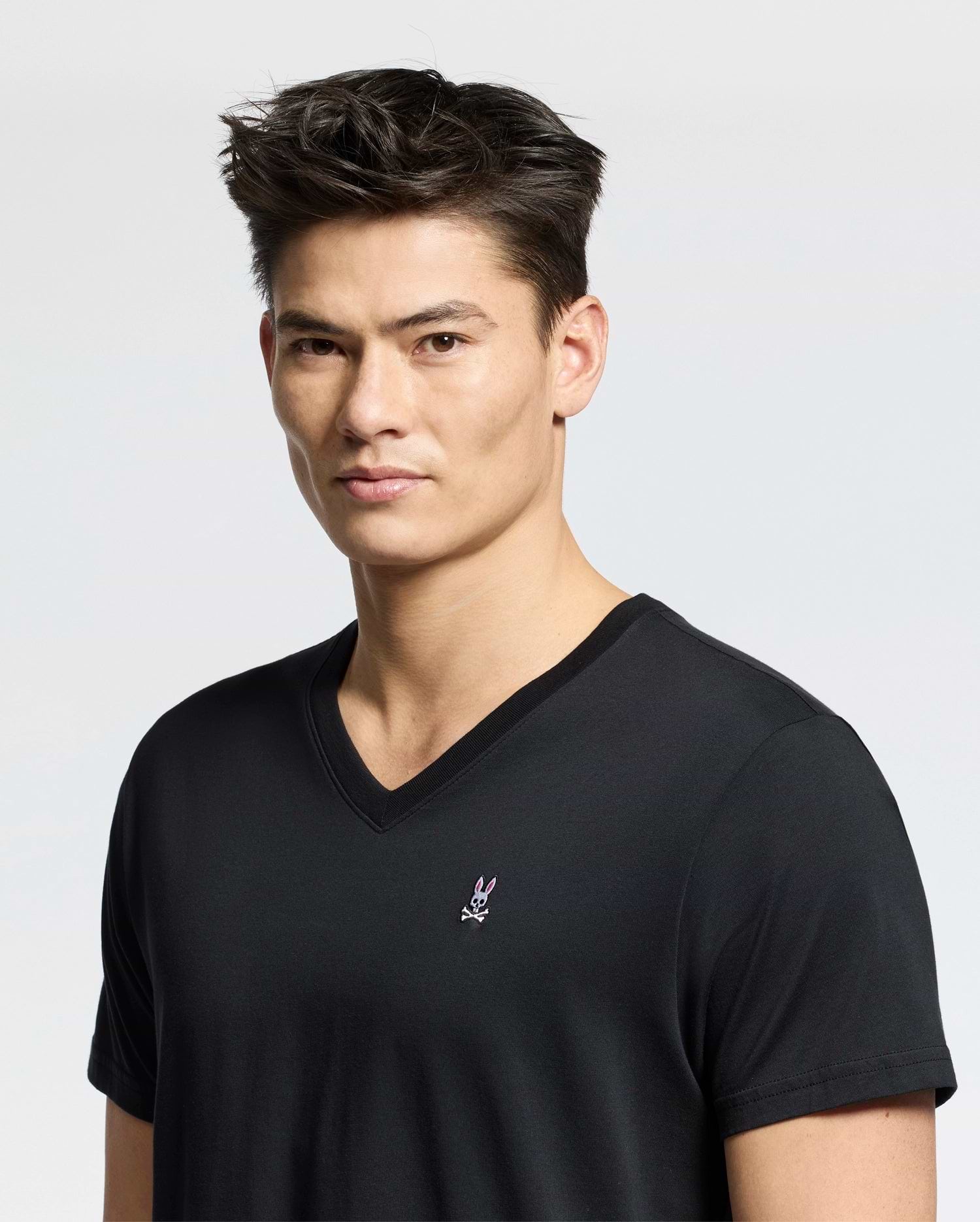 A person with short dark hair is wearing a Psycho Bunny MENS CLASSIC V NECK TEE, B6U100ARPC, featuring a small embroidered emblem on the chest. They are standing against a plain background and looking directly at the camera.