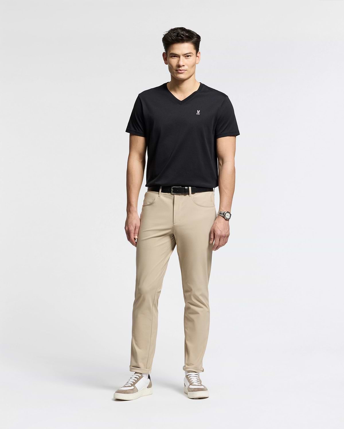 A man exudes confidence in a sleek black Psycho Bunny MENS CLASSIC V NECK TEE - B6U100ARPC, crafted from soft Pima cotton. Paired with beige pants and white sneakers, he embodies effortless style, his wrist adorned with a watch as he gazes forward against a plain background.