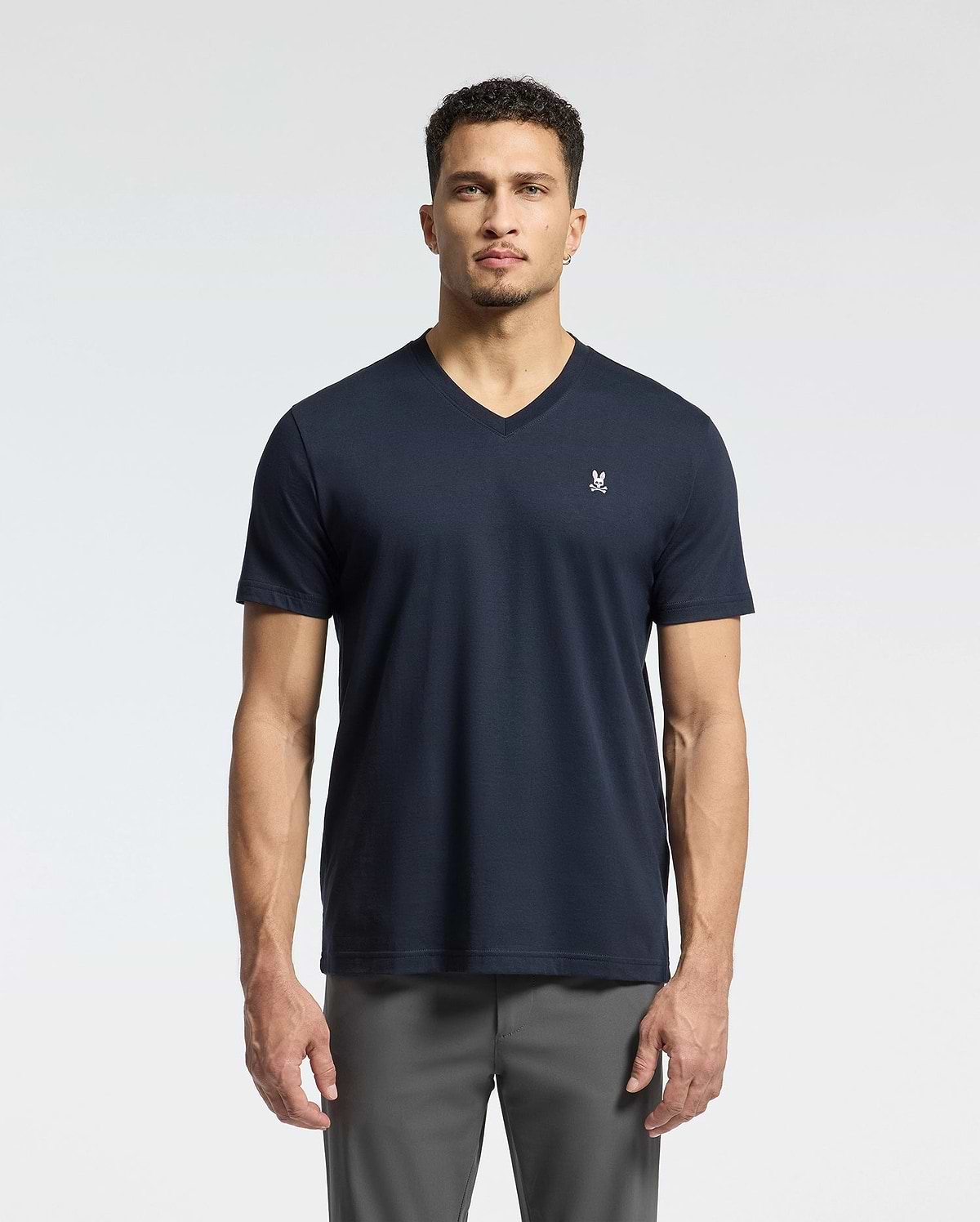Against a plain backdrop, a person with short curly hair and a neutral expression faces the camera. They're wearing Psycho Bunny's MENS CLASSIC V NECK TEE in dark Pima cotton, featuring a small white logo on the left chest, paired with gray pants.