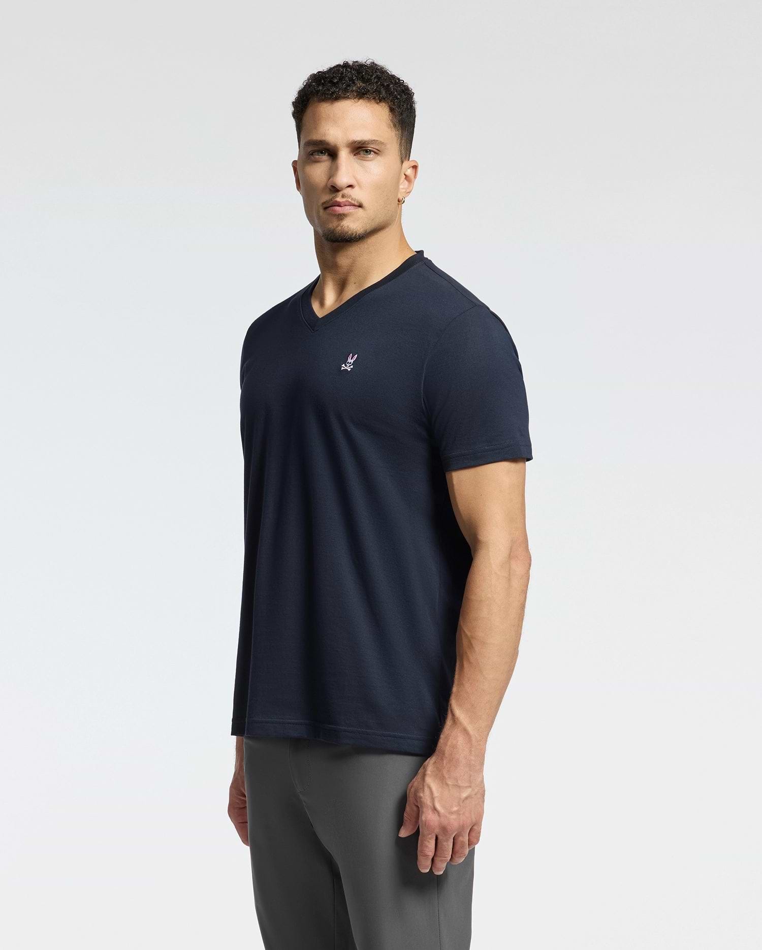 A person with short curly hair is wearing a dark blue Psycho Bunny MENS CLASSIC V NECK TEE - B6U100ARPC made from Pima cotton and gray pants, standing against a plain, light-colored background.