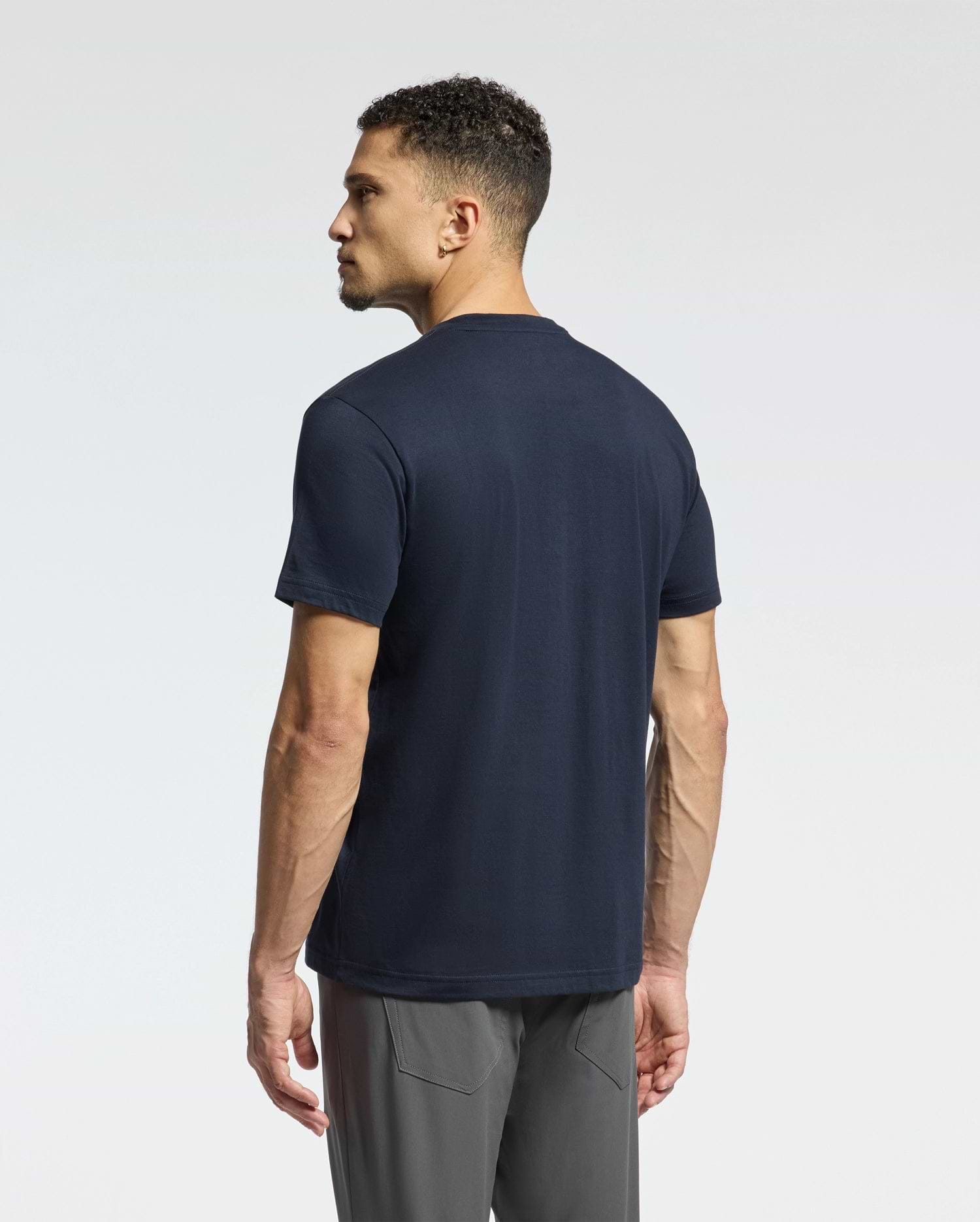 The man with short curly hair stands facing away, wearing a Psycho Bunny MENS CLASSIC V NECK TEE - B6U100ARPC in navy blue and gray pants against a plain light background.