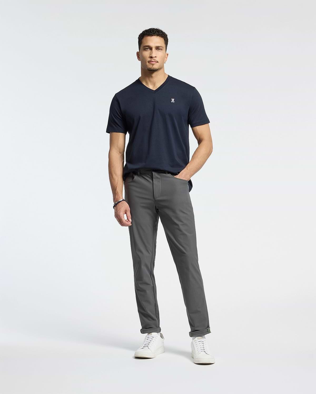A man stands against a white background wearing gray pants and the Psycho Bunny MENS CLASSIC V NECK TEE - B6U100ARPC in dark navy. Made of Pima cotton, it pairs well with his short curly hair and white sneakers. His right hand is in his pocket, with his left arm by his side.